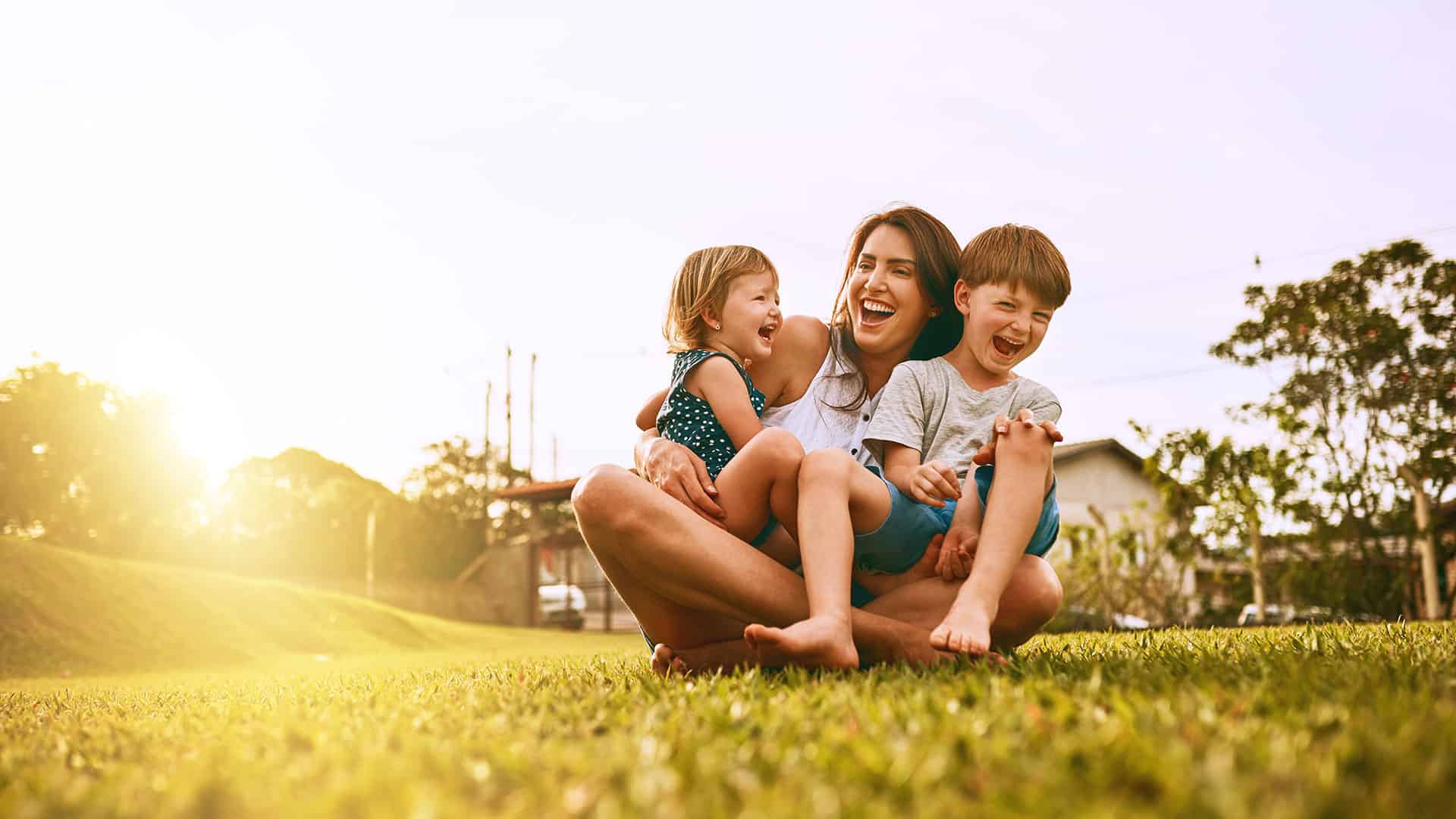 Mother with two children sitting in her lap on the grass with the sun behind her thinking about mother's day at shady maple.